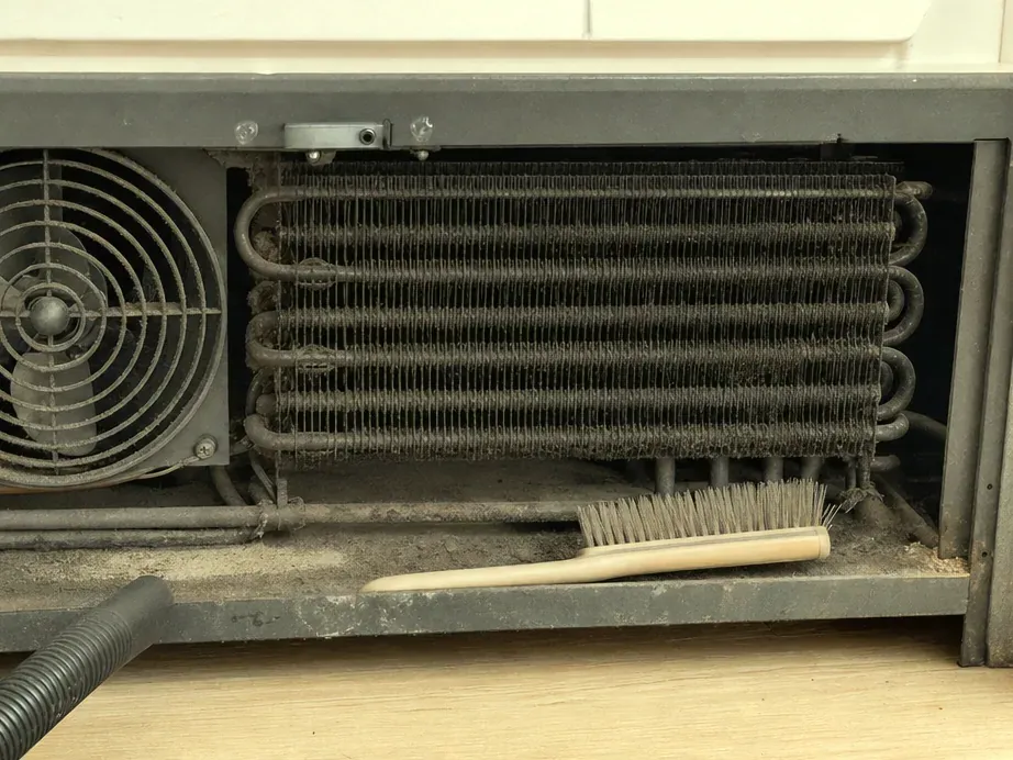 Condenser coil and fan with a long-bristle brush and a vacuum hose resting below.