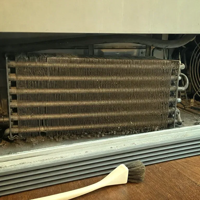 Dusty condenser coil at the base of a refrigerator, a coil brush lying on the floor in front.