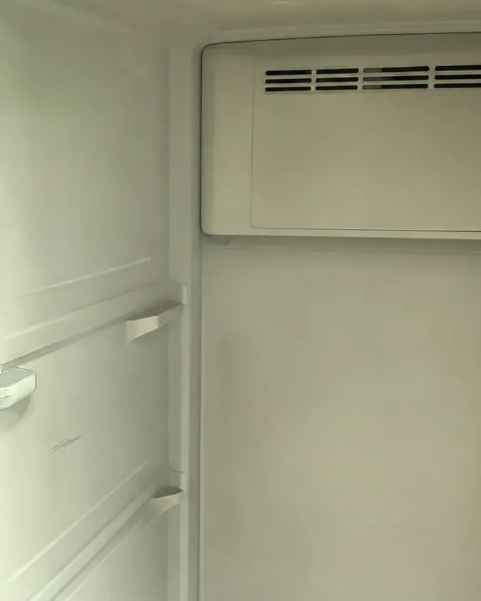 Empty white refrigerator compartment, a slotted vent cover high on the back wall and shelf supports along the side wall.